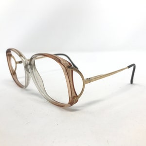 May include: Vintage eyeglasses with a clear and brown gradient frame and gold-tone metal arms. The glasses have a rounded, oversized shape. The earpieces are black. The glasses are set against a white background.