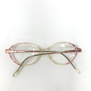 May include: Clear oval eyeglasses with a floral pattern on the frames. The frames have a pink, red, and white design. The arms are silver-toned metal.