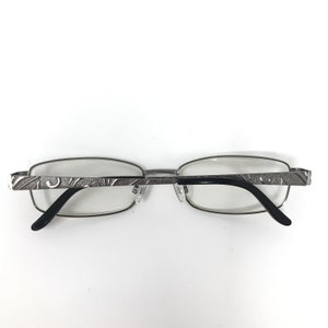 May include: Silver-framed eyeglasses with a rectangular lens shape. The temples feature an etched design. The earpieces are black. The glasses are set against a white background.