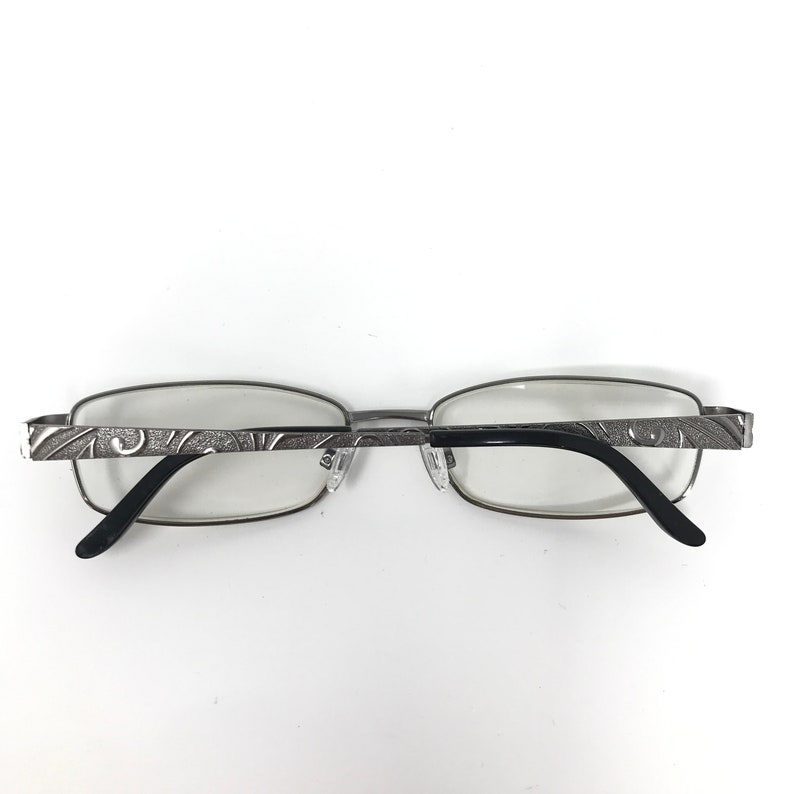 May include: Silver-framed eyeglasses with a rectangular lens shape. The temples feature an etched design. The earpieces are black. The glasses are set against a white background.