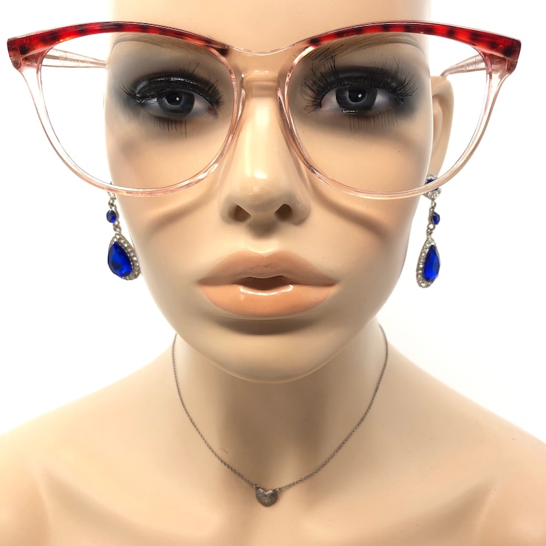 May include: Clear-framed eyeglasses with red and brown patterned upper rims. The mannequin wears blue teardrop earrings with silver accents and a small silver heart pendant necklace. The glasses are a cat-eye style.