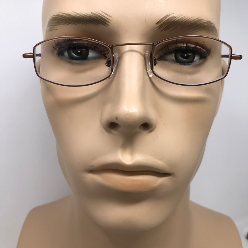 May include: Close-up of eyeglasses with a thin, bronze-colored metal frame. The rectangular lenses are clear, and the nose pads are transparent. The glasses are displayed on a mannequin head against a white background. The overall style is minimalist and modern.