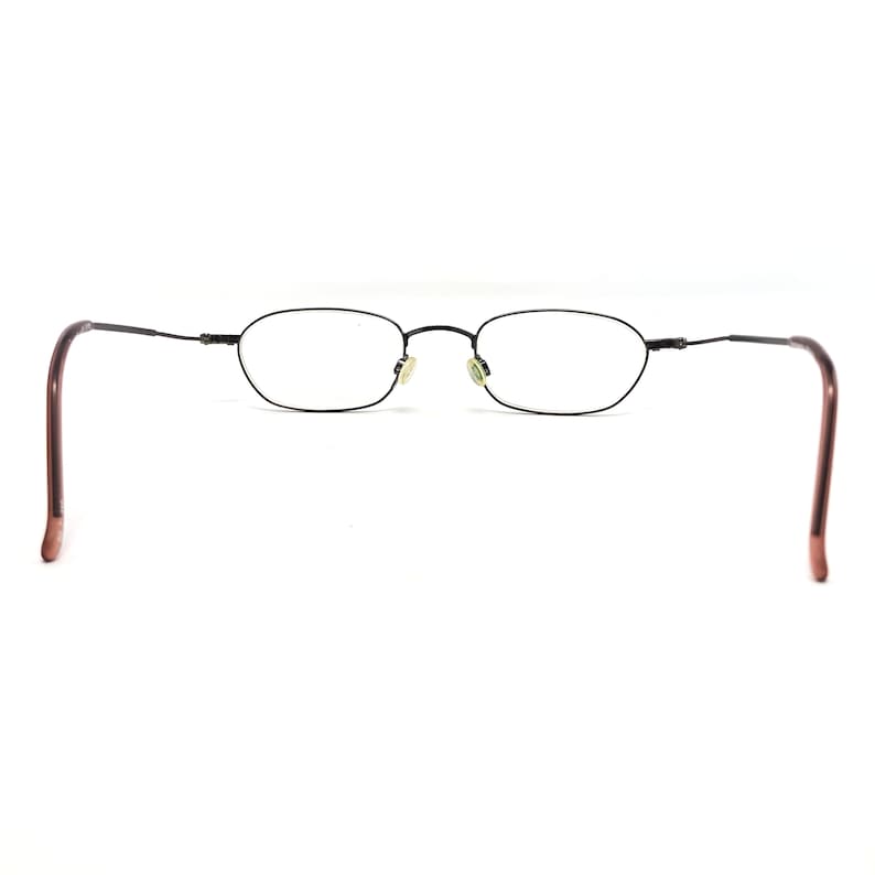 Puede incluir: Gafas ovaladas negras con montura met&aacute;lica fina y patillas marrones. Las lentes transparentes destacan sobre un fondo blanco. Dise&ntilde;o minimalista y cl&aacute;sico.