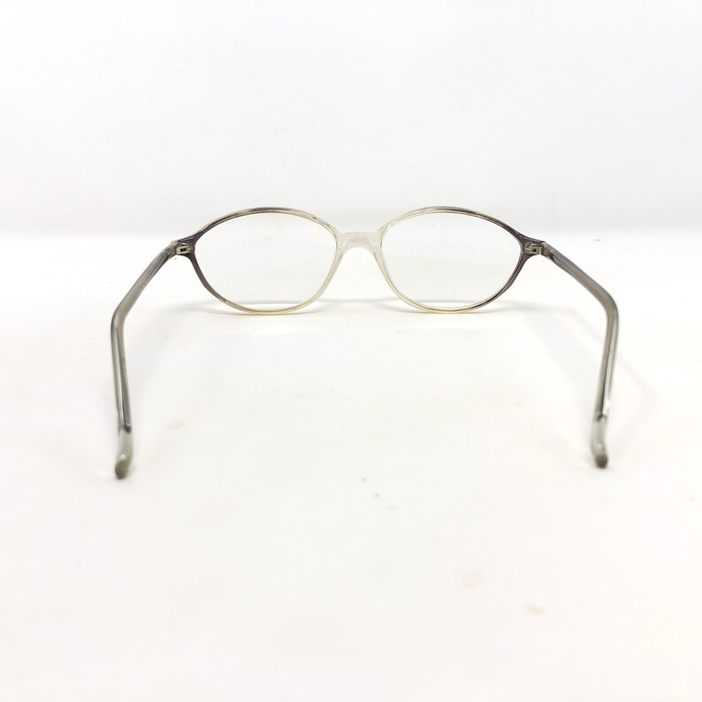 May include: Oval-shaped eyeglasses with a gradient frame, transitioning from a dark gray to a clear, translucent color. The temples are a solid gray. The glasses are set against a white background.