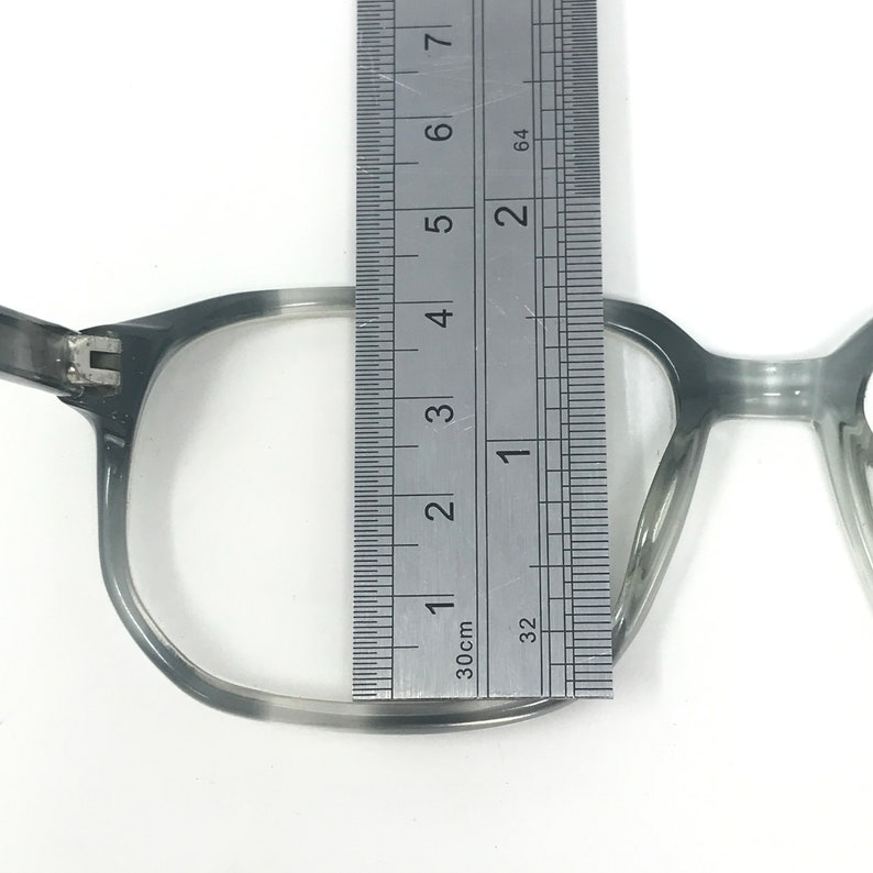 Puede incluir: Gafas grises con una regla que mide el ancho de la lente. El ancho de la lente es de aproximadamente 3,8 cm. Las gafas tienen forma rectangular y un color gris degradado.