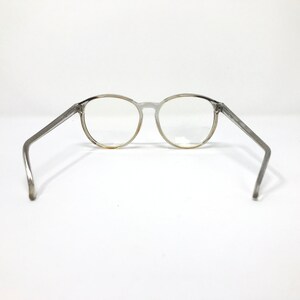 Puede incluir: Gafas transparentes de montura redonda con patillas grises. Las gafas se colocan sobre un fondo blanco, mostrando sus lentes transparentes y los detalles sutiles del dise&ntilde;o de la montura. Las patillas de las gafas est&aacute;n anguladas hacia afuera.