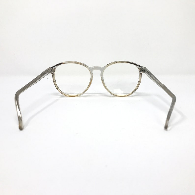 Puede incluir: Gafas transparentes de montura redonda con patillas grises. Las gafas se colocan sobre un fondo blanco, mostrando sus lentes transparentes y los detalles sutiles del dise&ntilde;o de la montura. Las patillas de las gafas est&aacute;n anguladas hacia afuera.