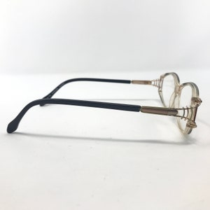 Puede incluir: Un par de gafas con montura transparente y patillas negras. Las lentes son ovaladas y la montura tiene detalles dorados. Las gafas est&aacute;n sobre un fondo blanco.