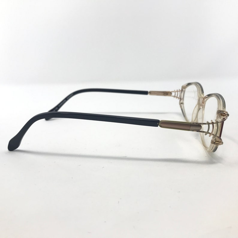 Puede incluir: Un par de gafas con montura transparente y patillas negras. Las lentes son ovaladas y la montura tiene detalles dorados. Las gafas est&aacute;n sobre un fondo blanco.