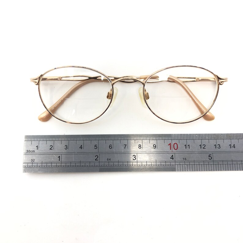 K&ouml;nnte beinhalten: Goldfarbene, oval geformte Brille mit klaren Gl&auml;sern. Die Brille hat einen filigranen Rahmen und B&uuml;gel. Ein Metalllineal mit Zentimetermarkierungen liegt darunter, zur Messung.