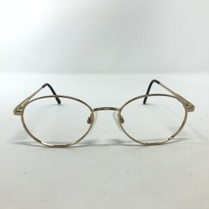 Pu&ograve; includere: Occhiali ovali color oro con lenti trasparenti. Le montature hanno un design sottile, con aste nere e naselli. Gli occhiali sono posti su uno sfondo bianco.