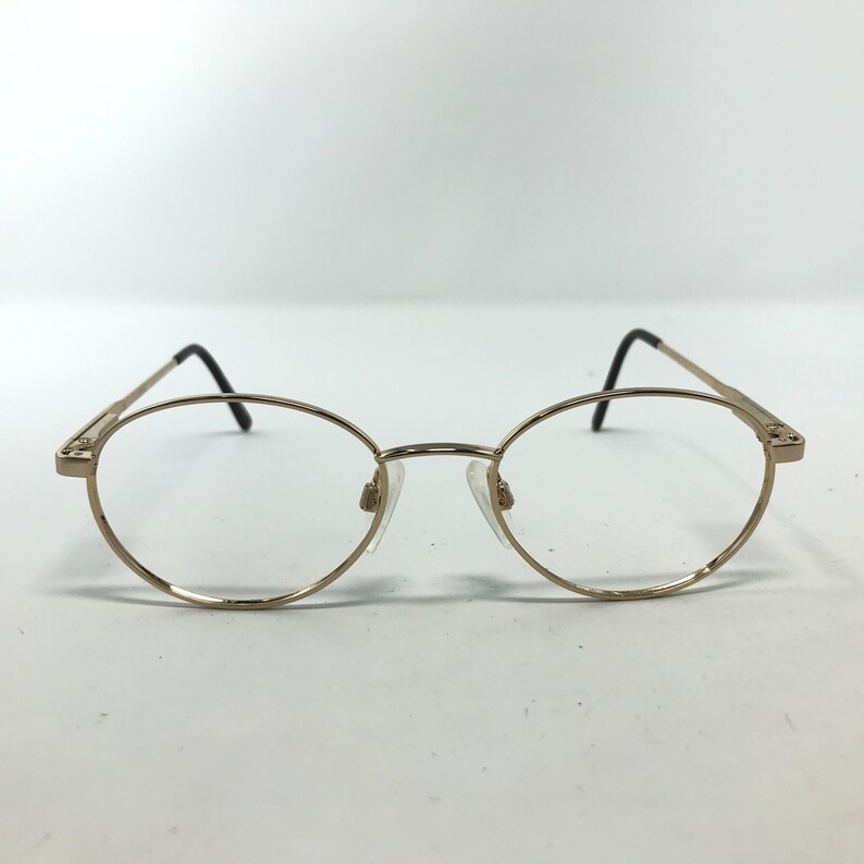 Pu&ograve; includere: Occhiali ovali color oro con lenti trasparenti. Le montature hanno un design sottile, con aste nere e naselli. Gli occhiali sono posti su uno sfondo bianco.