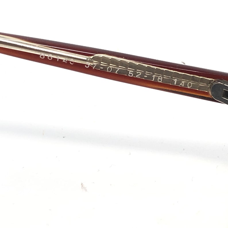 Puede incluir: Primer plano de una montura de gafas marr&oacute;n y beige. La montura tiene los n&uacute;meros "37-07 52-18 140" impresos en la patilla. La montura est&aacute; en &aacute;ngulo diagonal sobre un fondo blanco.