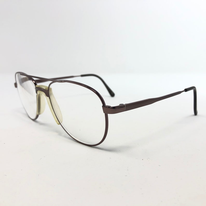 May include: A pair of aviator-style eyeglasses with a dark brown metal frame and clear lenses. The glasses have a double bridge and a classic teardrop shape. The nose pads are a light beige color.