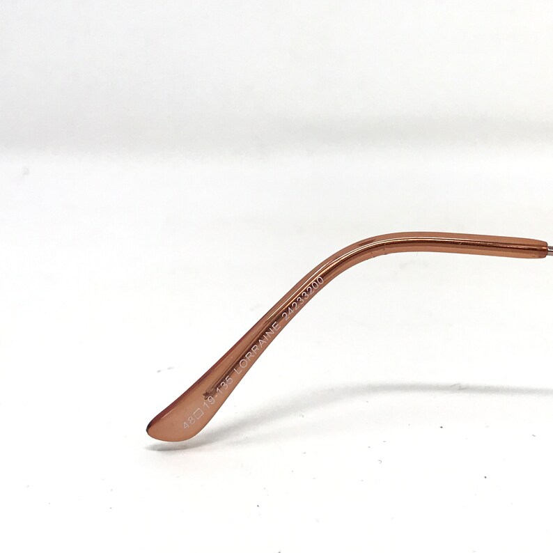 May include: Close-up of a rose gold eyeglasses arm. The arm features the text "48 19-135 LORRAINE 2423200" in black. The glasses arm is curved and has a smooth, metallic finish.