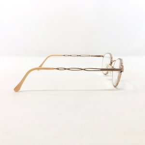 May include: Gold-toned eyeglasses with a round frame and a decorative, interwoven design on the temples. The glasses have a classic, elegant style, suitable for everyday wear. The lenses are clear.