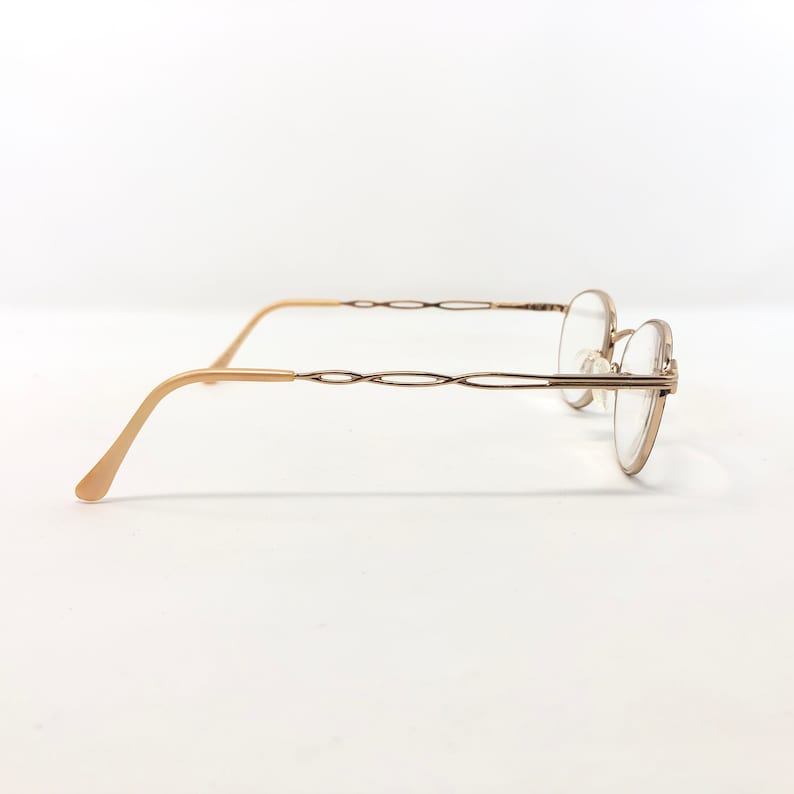 May include: Gold-toned eyeglasses with a round frame and a decorative, interwoven design on the temples. The glasses have a classic, elegant style, suitable for everyday wear. The lenses are clear.