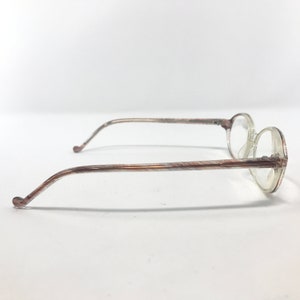 May include: A pair of vintage spectacles featuring clear, round lenses and slender, patterned arms. The frames are a light brown hue with a delicate, textured pattern. The glasses are set against a plain white backdrop.