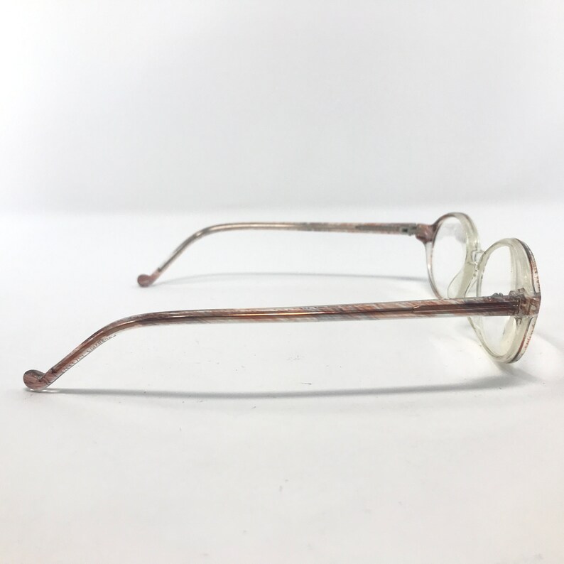 May include: A pair of vintage spectacles featuring clear, round lenses and slender, patterned arms. The frames are a light brown hue with a delicate, textured pattern. The glasses are set against a plain white backdrop.