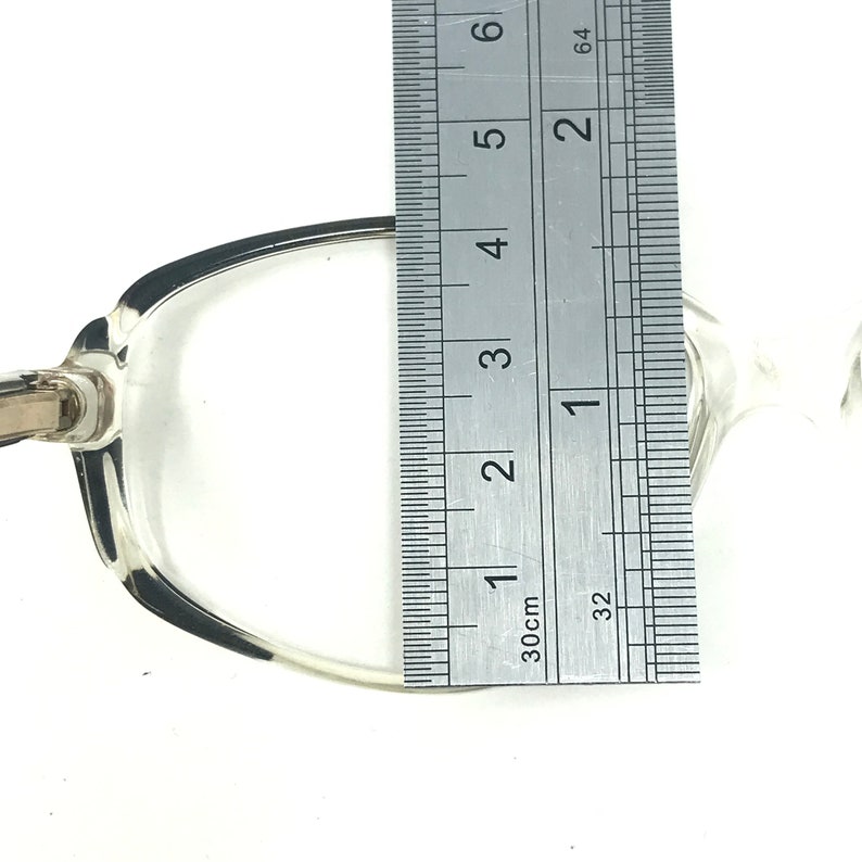 May include: Clear eyeglasses with a silver-coloured frame are measured with a ruler. The frame is approximately 10 cm wide. The glasses are set against a white background.