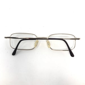 May include: Silver-framed eyeglasses with rectangular lenses and black temples. The glasses have clear lenses and nose pads. The frame is a thin, silver metal. The temples curve downward to end in black tips.
