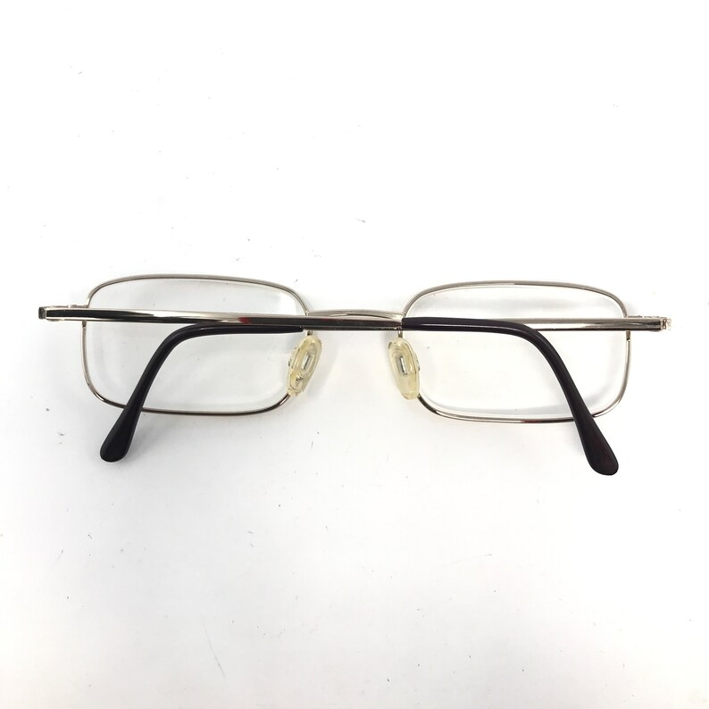 May include: Silver-framed eyeglasses with rectangular lenses and black temples. The glasses have clear lenses and nose pads. The frame is a thin, silver metal. The temples curve downward to end in black tips.