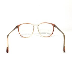 May include: Vintage eyeglasses with a gradient frame, transitioning from a light peach color to clear. The arms are a silver-tone metal with peach-colored tips. The lenses are clear, and the glasses are set against a white background.