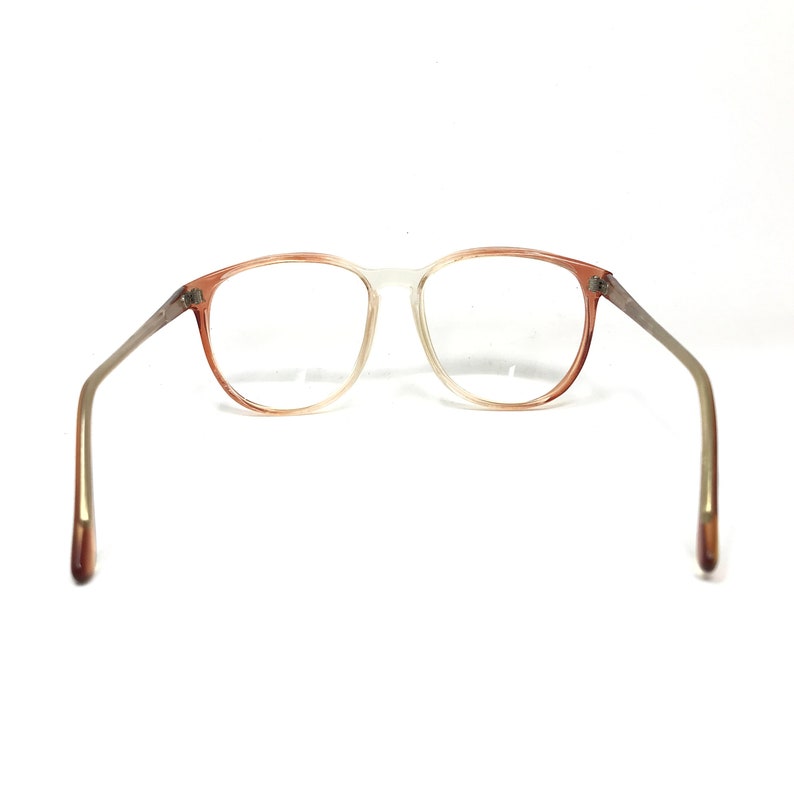 May include: Vintage eyeglasses with a gradient frame, transitioning from a light peach color to clear. The arms are a silver-tone metal with peach-colored tips. The lenses are clear, and the glasses are set against a white background.