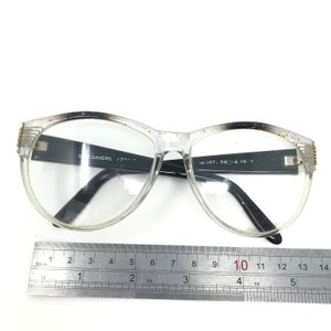 May include: Clear-framed eyeglasses with black temples and a speckled design along the top edges. The glasses have a rectangular shape and are positioned on a white surface. The text "Specsavers 17..." and "10 587, 58 16 YW 7" is visible on the frame.