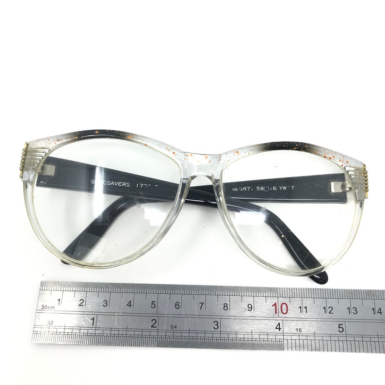 May include: Clear-framed eyeglasses with black temples and a speckled design along the top edges. The glasses have a rectangular shape and are positioned on a white surface. The text "Specsavers 17..." and "10 587, 58 16 YW 7" is visible on the frame.