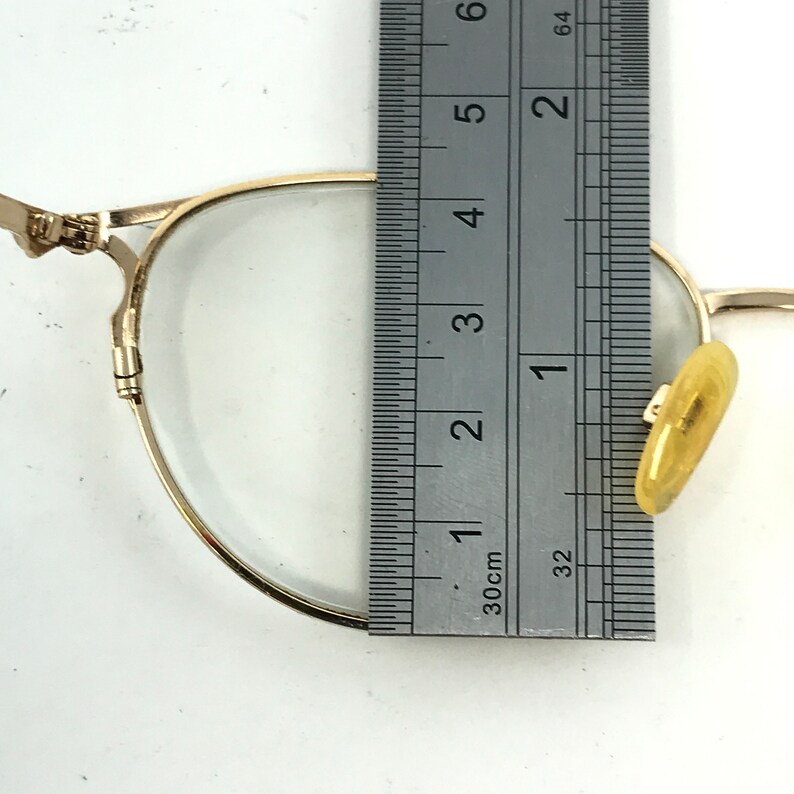 Pu&ograve; includere: Un paio di occhiali dorati con una lente trasparente e un'asta in plastica gialla. Gli occhiali sono misurati con un righello, mostrando che l'asta misura circa 3,8 cm di lunghezza.