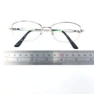 May include: Silver-rimmed eyeglasses with clear lenses, featuring a half-frame design. The glasses have black temples and are positioned on a white surface next to a ruler. The ruler shows measurements in inches and centimeters.