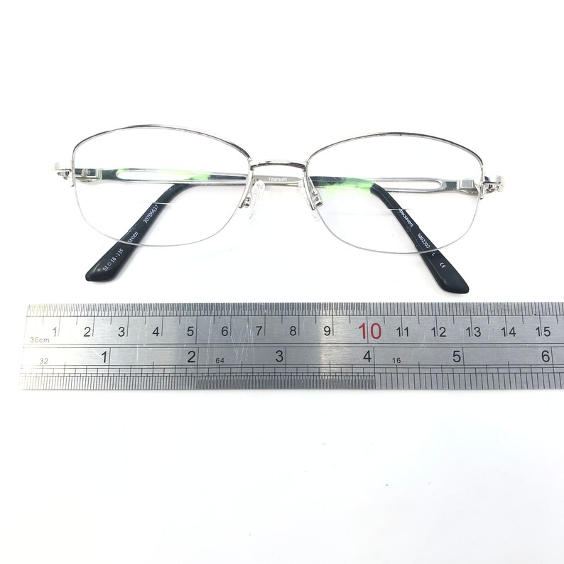 May include: Silver-rimmed eyeglasses with clear lenses, featuring a half-frame design. The glasses have black temples and are positioned on a white surface next to a ruler. The ruler shows measurements in inches and centimeters.