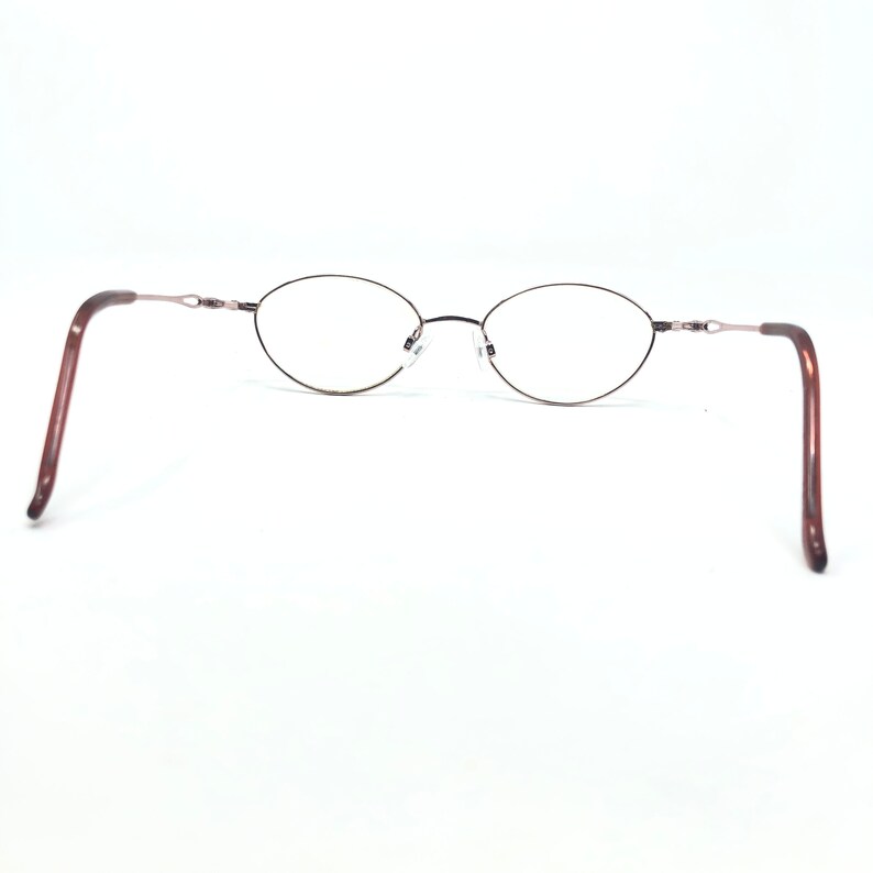 K&ouml;nnte beinhalten: Oval geformte Brille mit einem d&uuml;nnen, dunkelbraunen Metallrahmen. Die B&uuml;gel sind dunkler braun mit Schildpattmuster. Die Gl&auml;ser sind klar, und der Nasensteg ist silberfarben. Die Brille ist vor wei&szlig;em Hintergrund.