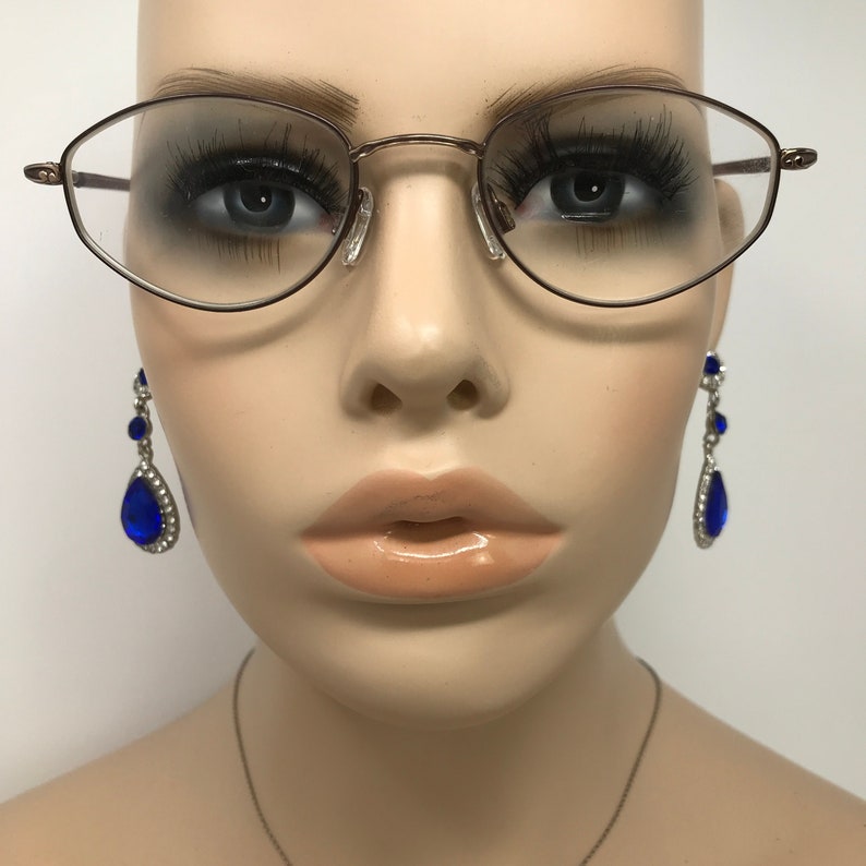 Puede incluir: Un busto de maniqu&iacute; que muestra gafas y pendientes. Las gafas tienen una montura fina de metal marr&oacute;n con forma ovalada. Los pendientes son plateados con gemas azules en forma de l&aacute;grima y pedrer&iacute;a transparente. El maniqu&iacute; tiene pesta&ntilde;as largas y un color de labios neutro.