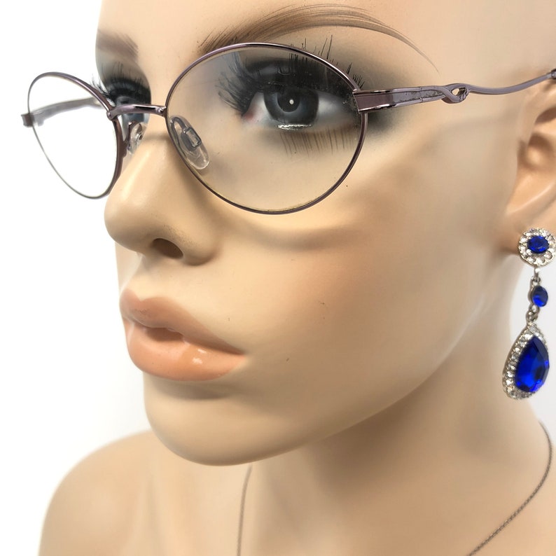 Peut inclure: Gros plan d'un mannequin portant des lunettes ovales &agrave; monture violette et des boucles d'oreilles pendantes en pierres pr&eacute;cieuses bleues. Les boucles d'oreilles ont un sertissage argent&eacute; et une forme de goutte d'eau. Les lunettes ont une monture d&eacute;licate en fil de fer.
