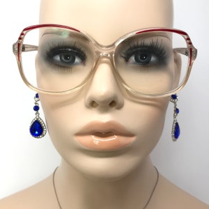 May include: Clear-framed eyeglasses with red and white accents on the upper rims. The glasses are displayed on a mannequin with long, dark eyelashes and a pair of blue teardrop earrings with silver settings.