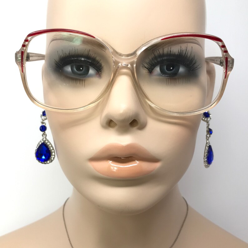 May include: Clear-framed eyeglasses with red and white accents on the upper rims. The glasses are displayed on a mannequin with long, dark eyelashes and a pair of blue teardrop earrings with silver settings.