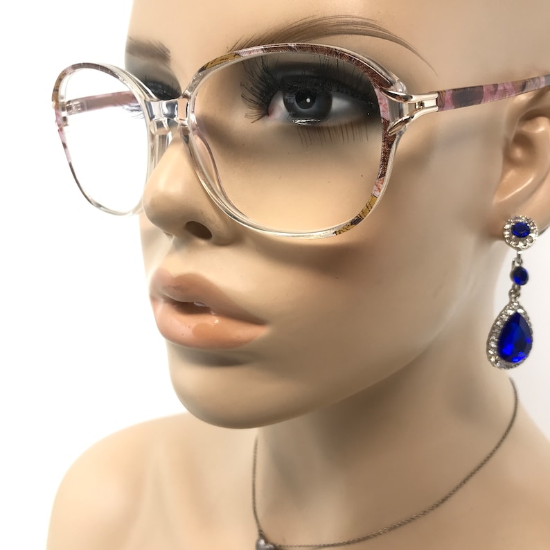 May include: Clear-framed eyeglasses with a floral pattern on the temples. The glasses are paired with blue teardrop earrings with silver accents. A mannequin head is wearing the glasses and earrings.
