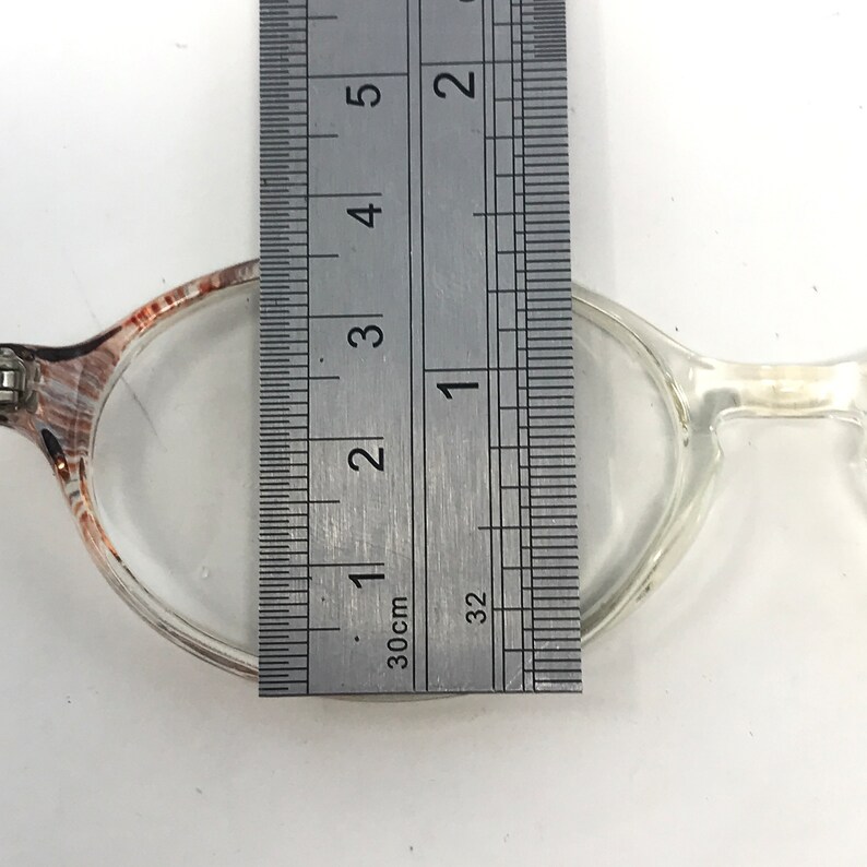 May include: Clear eyeglasses with a ruler measuring the frame's width. The ruler shows the frame is approximately 2.5 cm wide. The glasses have a clear frame with a patterned brown and white detail on the upper rim.
