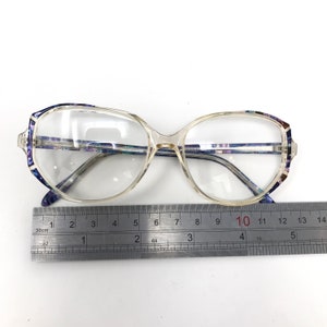 Puede incluir: Gafas transparentes con montura rectangular y detalles en azul y morado estampados en las patillas. Una regla met&aacute;lica se coloca debajo, midiendo aproximadamente 13 cm de ancho.