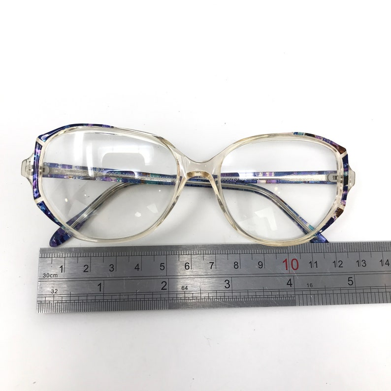 Puede incluir: Gafas transparentes con montura rectangular y detalles en azul y morado estampados en las patillas. Una regla met&aacute;lica se coloca debajo, midiendo aproximadamente 13 cm de ancho.