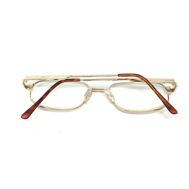 May include: Gold-tone metal eyeglasses with brown plastic arms. The glasses have a rectangular shape and are designed for reading.