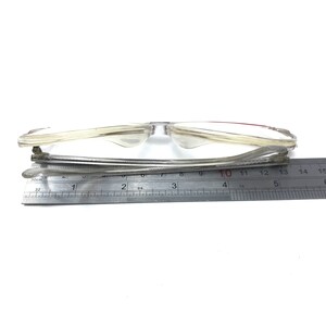 Pu&ograve; includere: Occhiali con montatura in plastica trasparente con un accento rosso sul ponte del naso. Gli occhiali sono misurati con un righello, mostrando una larghezza di circa 10 centimetri.
