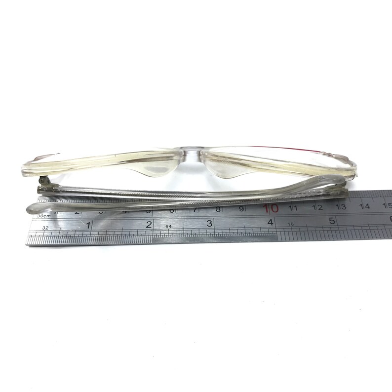 Pu&ograve; includere: Occhiali con montatura in plastica trasparente con un accento rosso sul ponte del naso. Gli occhiali sono misurati con un righello, mostrando una larghezza di circa 10 centimetri.