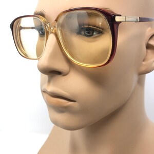 May include: Vintage eyeglasses with a square frame. The frames are a combination of brown and clear plastic with gold-colored metal accents on the temples. The lenses have a yellow tint. The glasses are displayed on a mannequin head.