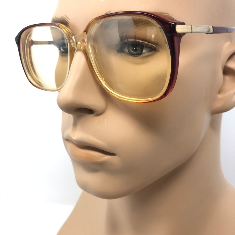 May include: Vintage eyeglasses with a square frame. The frames are a combination of brown and clear plastic with gold-colored metal accents on the temples. The lenses have a yellow tint. The glasses are displayed on a mannequin head.