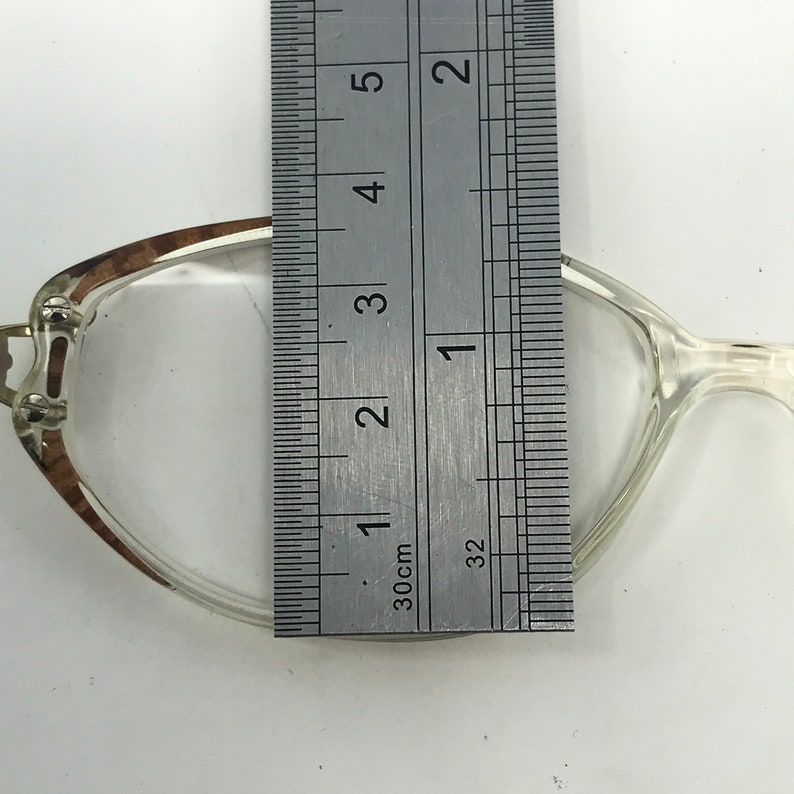 K&ouml;nnte beinhalten: Transparente Brille mit braun-silbernem Rahmen, gemessen mit einem Lineal. Das Lineal zeigt, dass die Brille etwa 2.5 cm breit ist. Auf einer wei&szlig;en Oberfl&auml;che.