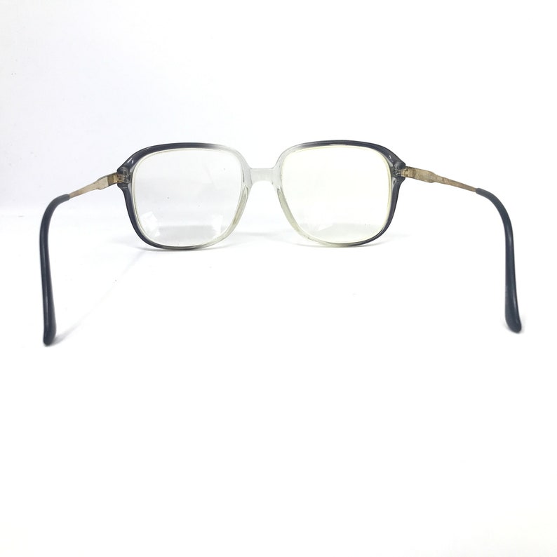 Pu&ograve; includere: Occhiali vintage con montatura quadrata con montatura in plastica grigia sfumata e trasparente. Le aste sono in metallo dorato e i terminali delle aste sono neri. Gli occhiali sono su sfondo bianco.