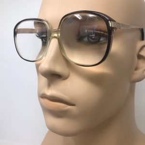 May include: Vintage eyeglasses with a brown and clear frame. The glasses have a square shape and a gold-colored metal arm. The lenses are clear and reflect the surrounding environment.